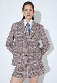 Geruite blazer in beige met rode en groene accenten, voorzien van brede revers en twee knopen, gecombineerd met een bijpassende geplooide minirok.