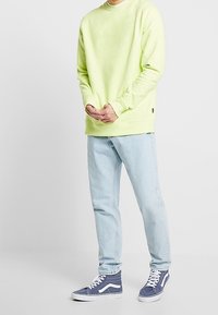 Jasnozielona bluza crewneck z prążkowanymi mankietami, zestawiona z jasnoniebieskimi dżinsami i niebieskimi trampkami z białymi akcentami. Swobodny look streetwear.