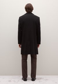 Abrigo de lana negro con un diseño entallado, abertura única en la parte trasera. Pantalones marrones de tela texturizada, zapatos negros pulidos.