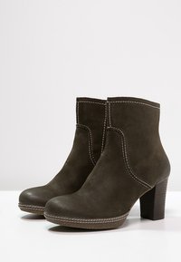 Botas de ante marrón oscuro hasta el tobillo con tacones altos gruesos y costuras blancas visibles a lo largo de los bordes, colocadas sobre una superficie blanca.