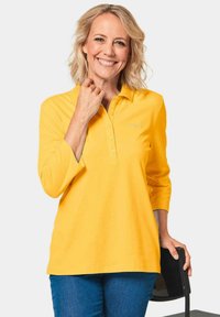 Geel longsleeve polo shirt met een drie knoopjes sluiting, lichtgewicht stof en subtiele borduursel, gecombineerd met blauwe spijkerbroeken.