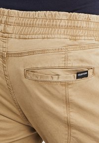 Beige broek met elastische tailleband, achterzak met een label "YOURTURN" en zichtbare stikdetails. Glad van textuur.