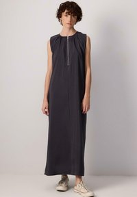 Abito senza maniche, lungo fino al pavimento, in tessuto grigio scuro con una leggera lucentezza. Presenta una zip frontale e arricciature all'area del collo. Indossato con sneakers leggere.