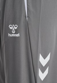 Szara tkanina z białym logo pszczoły hummel i dwoma skierowanymi w dół pasami w kształcie szewrona, prawdopodobnie materiał odzieży sportowej z widocznym detalem szwu.