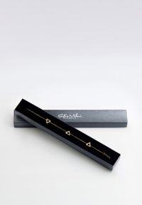 Guld geometriskt armband med triangulära berlocker, visat i en svart låda med en mjuk insida och silverlogotyp på locket.