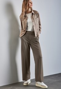 Beige, long-sleeve shirt jacket with pockets, paired with loose, dark green trousers and white sneakers, set against a light wall.