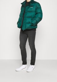 Chaqueta acolchada verde con cremallera frontal, cuello alto y costuras visibles. Lleva puesta una camisa negra, combinada con vaqueros oscuros y zapatillas blancas.