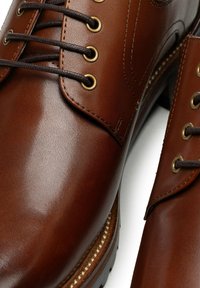 Brązowe skórzane buty o gładkim wykończeniu, z metalowymi przelotkami, brązowymi sznurówkami oraz kontrastującą jasnobrązową podeszwą z przeszyciami.