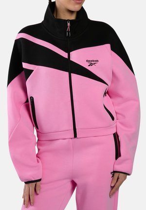 Vrouw draagt een roze-zwart Reebok trainingspak met een ritsjack en bijpassende broek, staand met haar handen ontspannen langs haar zij.