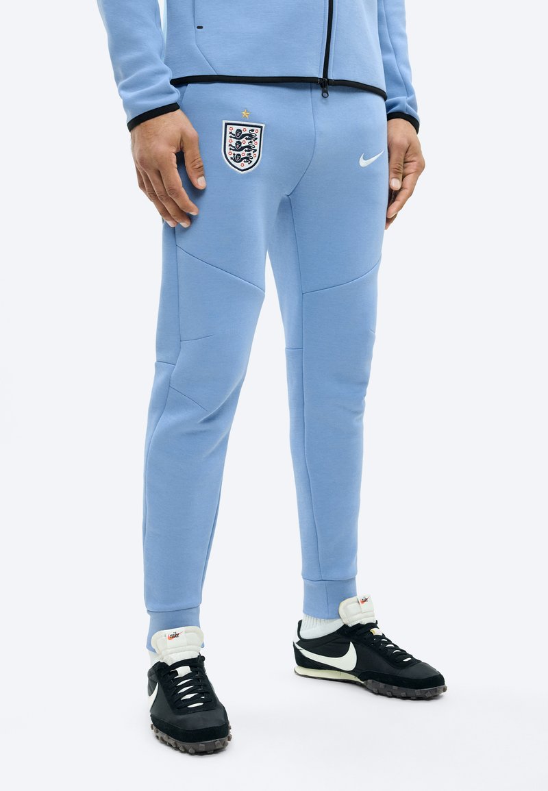 Άτομο που φοράει ανοιχτό μπλε αθλητική φόρμα Αγγλίας με το λογότυπο της Nike και μαύρα και λευκά αθλητικά παπούτσια Nike, στέκεται σε λευκή επιφάνεια.