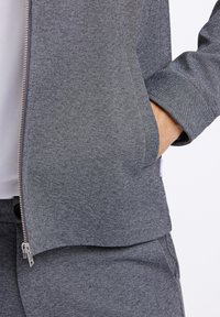 Grå zip-up jacka med strukturerat tyg, inkluderar sidofickor, muddar med knappdetalj, matchas med grå byxor.