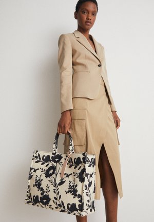 Kvinde iført beige blazer og nederdel med frontslids, der holder en stor cremefarvet shopper med sort blomsterprint og håndtag.