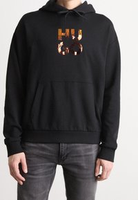 Sudadera negra hecha de tela suave que presenta un gran logo reflectante en cobre "HUGO" en la parte frontal y un bolsillo en la parte delantera. Incluye capucha ajustable.