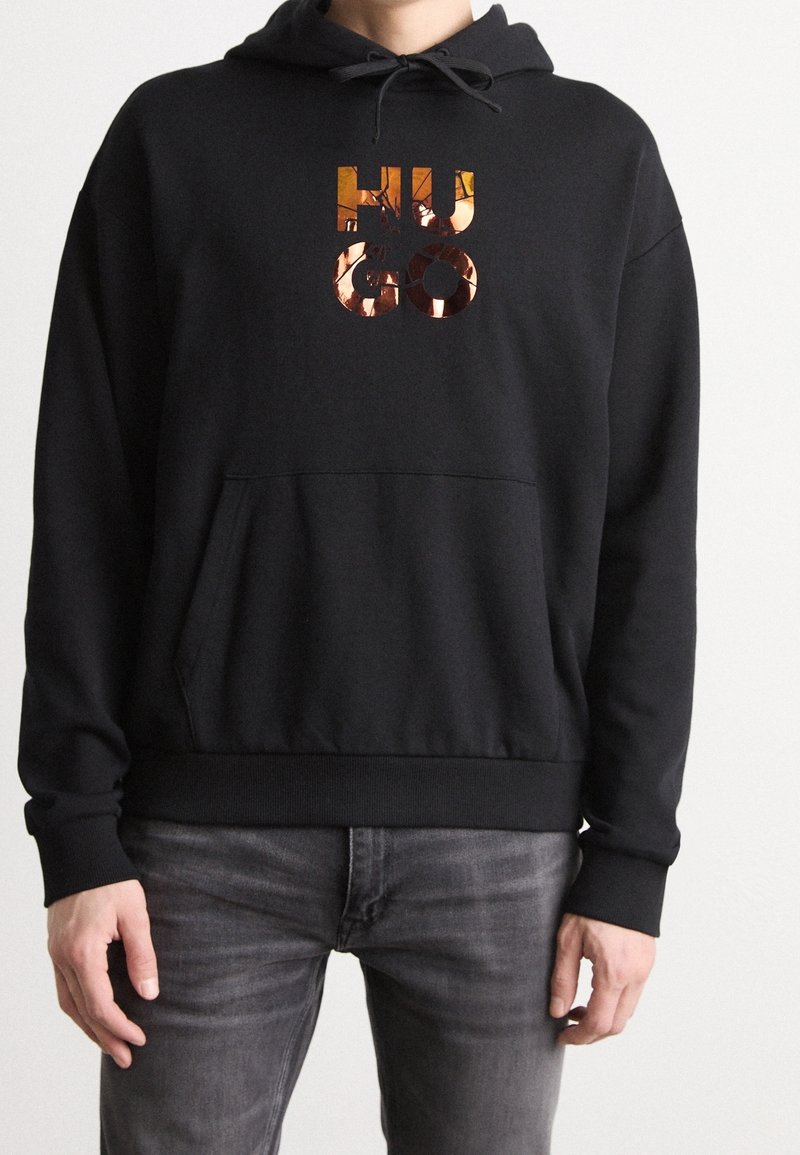 Sudadera negra hecha de tela suave que presenta un gran logo reflectante en cobre "HUGO" en la parte frontal y un bolsillo en la parte delantera. Incluye capucha ajustable.