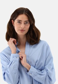 Hellblaues, übergroßes Hemd aus einem weichen Stoff. Hat einen V-Ausschnitt, lange Ärmel mit Knopfmanschetten und dezente Steppnähte.