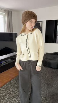 Beiger Strickjacke mit schwarzem Paspelier und Taschen, getragen über dunkler Hose. Das Modell trägt außerdem einen braunen Pelzhut und goldene Creolen.