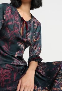 Blommönstrad klänning i mörkgrönt tyg, med en nyckelhålsurringning med knytning, långa ärmar med volangmuddar och en flödande silhuett.