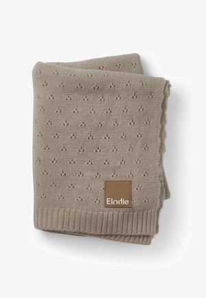 Knit blanket in beige with a textured pattern of small dots, scalloped edges, and a brown label featuring white text. Soft, warm material.