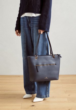 Person wearing navy sweater and wide-leg jeans holding black leather tote bag with gold zipper and white pointed shoes on wooden floor.