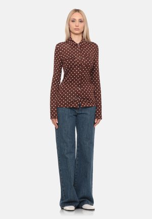 Donna con capelli biondi lisci indossa una camicia marrone con bottoni e pois bianchi, jeans blu a gamba larga e scarpe bianche, in piedi di fronte.