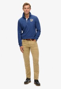 Blue long-sleeve polo with a quarter zip and embroidered logo, paired with khaki chinos and black shoes, featuring a braided brown belt.