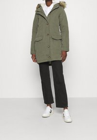 Olivgrön parkas med pälsfoder på huven, knapplås och två framfickor. Kombinerad med svarta byxor och vita sneakers.