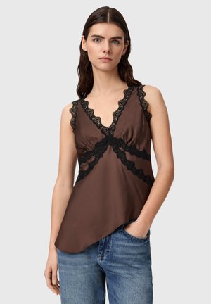 Kobieta ubraną w brązowy top bez rękawów z czarną koronkową lamówką i niebieskie dżinsy, stojąca z jedną ręką w kieszeni na jednolitym tle.