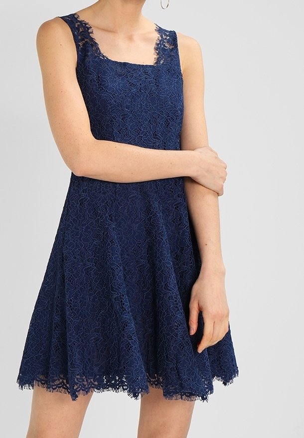 Robe sans manches en dentelle bleu marine avec un décolleté carré et une jupe évasée portée par une personne les bras croisés.