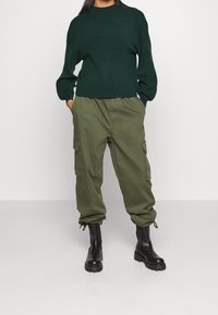 Tummanvihreä neulottu pitkähihainen pooloneule, yhdistetty oliivinvihreisiin cargo-housuihin, joissa on sivutaskut, sekä mustiin nilkkureihin, joissa on paksut pohjat.