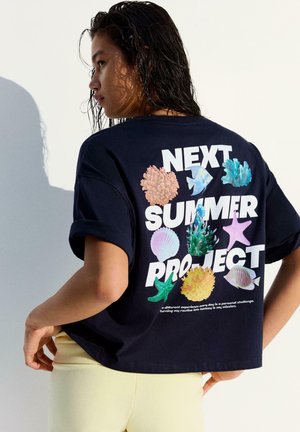 Giovane donna con capelli bagnati che indossa una maglietta nera con grafiche di vita marina e la scritta "Next Summer Project" sul retro.