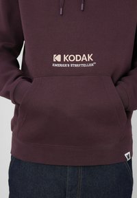 Personne portant un hoodie bordeaux avec le texte et le logo "Kodak America's Storyteller" à l'avant, les mains dans la poche kangourou, associé à un jean en denim foncé.