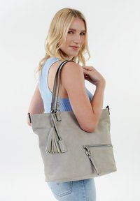 Gray faux leather tote bag with dual straps, zippered pocket, tassel detail, and metal hardware accents. Smooth texture and minimalist design.