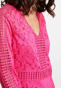 Top rosa con escote en V, tejido texturizado con patrones geométricos y detalles de calado a lo largo de las mangas y la cintura.