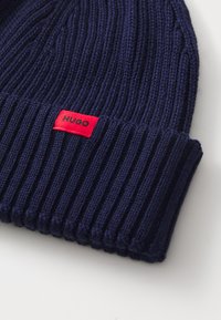 Gorro de punto en color navy con textura acanalada, que presenta una etiqueta roja con la inscripción "HUGO". El dobladillo es grueso y detallado, lo que realza su apariencia estructurada.