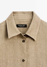 Jasnobrązowa koszula z kołnierzykiem, wykonana z teksturowanej tkaniny, z czarnymi guzikami, oznaczona etykietą "Massimo Dutti" oraz "Wyprodukowano w Portugalii".