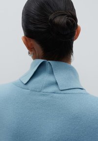 Frau mit dunklem Haar zu einem Dutt getragen, mit silbernen Creolen und einem hellblauen Strickpullover mit umgeschlagenem Kragen, von hinten betrachtet.