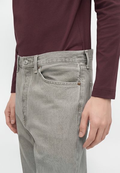 Personne portant un jean gris en denim et une chemise à manches longues rouge foncé, main reposant le long du corps, montrée de la taille à mi-cuisse.
