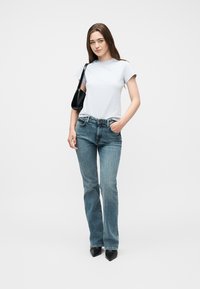 Νεαρή γυναίκα με μακριά καστανά μαλλιά που φοράει λευκό μπλουζάκι, μπλε τζιν, μαύρα μυτερά παπούτσια και μαύρη τσάντα ώμου, στέκεται σε λευκό φόντο.