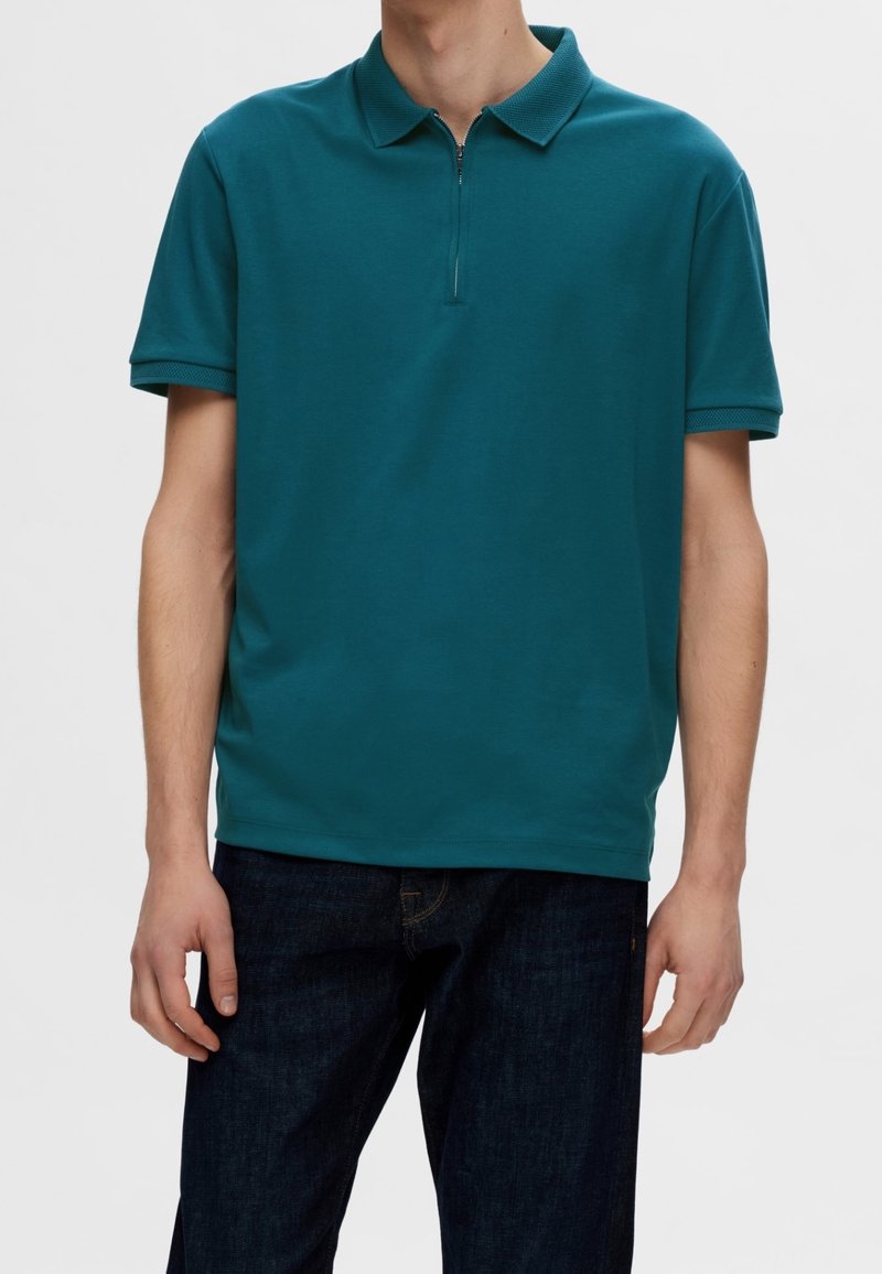 Polo à manches courtes couleur sarcelle avec col zippé porté par une personne, associé à un jean bleu foncé sur un fond uni.