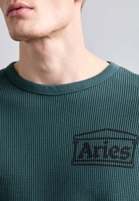 Green ribbed crewneck shirt featuring a dark gray logo "Aries" on the left side, crafted from soft textured material.