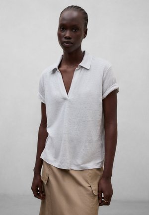 Frau mit geflochtenem Haar, die ein hellgraues kurzärmeliges Hemd mit Kragen und einen beigen Rock vor einem einfarbigen grauen Hintergrund trägt.