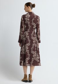 Langes kastanienbraunes Kleid mit weißem Paisleymuster, Kragen, langen Ärmeln und gebundenem Taillenzug. Dazu passen Wildleder-Ankle-Boots.