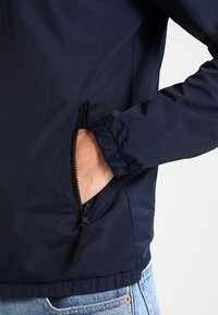 Námořnická modrá lehká bunda s kapsou na zip, elastickými manžetami a hladkým, mírně texturovaným materiálem.