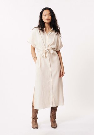 Robe boutonnière beige clair avec manches courtes, ceinture à la taille et fentes sur les côtes. Portée avec des bottes de cowboy marron. Tissu à texture lisse.