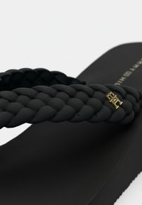 Μαύρο σαγιονάρι με πλεγμένους ιμάντες από ευλύγιστο υλικό, με χρυσό διακοσμητικό λογότυπο στον ιμάντα. Υφή σε μαύρη σόλα.