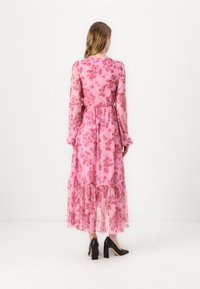 Robe longue rose en tissu translucide avec un motif floral, manches longues, ourlet à volants et taille ajustée. Associée à des chaussures à talons hauts noires.