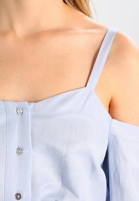 Camicia azzurra chiara a maniche corte con chiusura a bottoni, caratterizzata da una vestibilità rilassata e una morbida texture in cotone. Spalle scoperte e sottili spalline.
