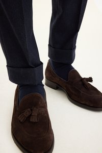 Bruna mockaläders loafers med tofsdekorationer, kombinerade med marinblå strumpklädda ben och mörka byxor, som visar uppvikta fållar och texturerat tyg.