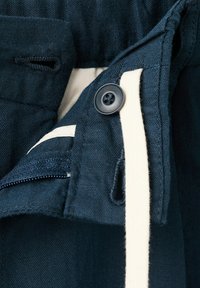 Donkerblauwe stof met een knoopsgat en een zwarte knop die een beige strook vastzet, naast een gedeeltelijk zichtbare ritssluiting op een kledingstuk.