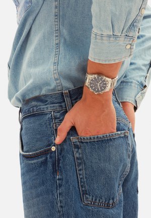 Person iført lyseblå cowboyskjorte og blå jeans, hånd i baglomme, iført sølvfarvet armbåndsur med mørk urskive.